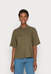 Camisa de manga curta, verde oliva, com fecho de botões à frente, colarinho e um único bolso no peito. Feita de um tecido liso e leve.