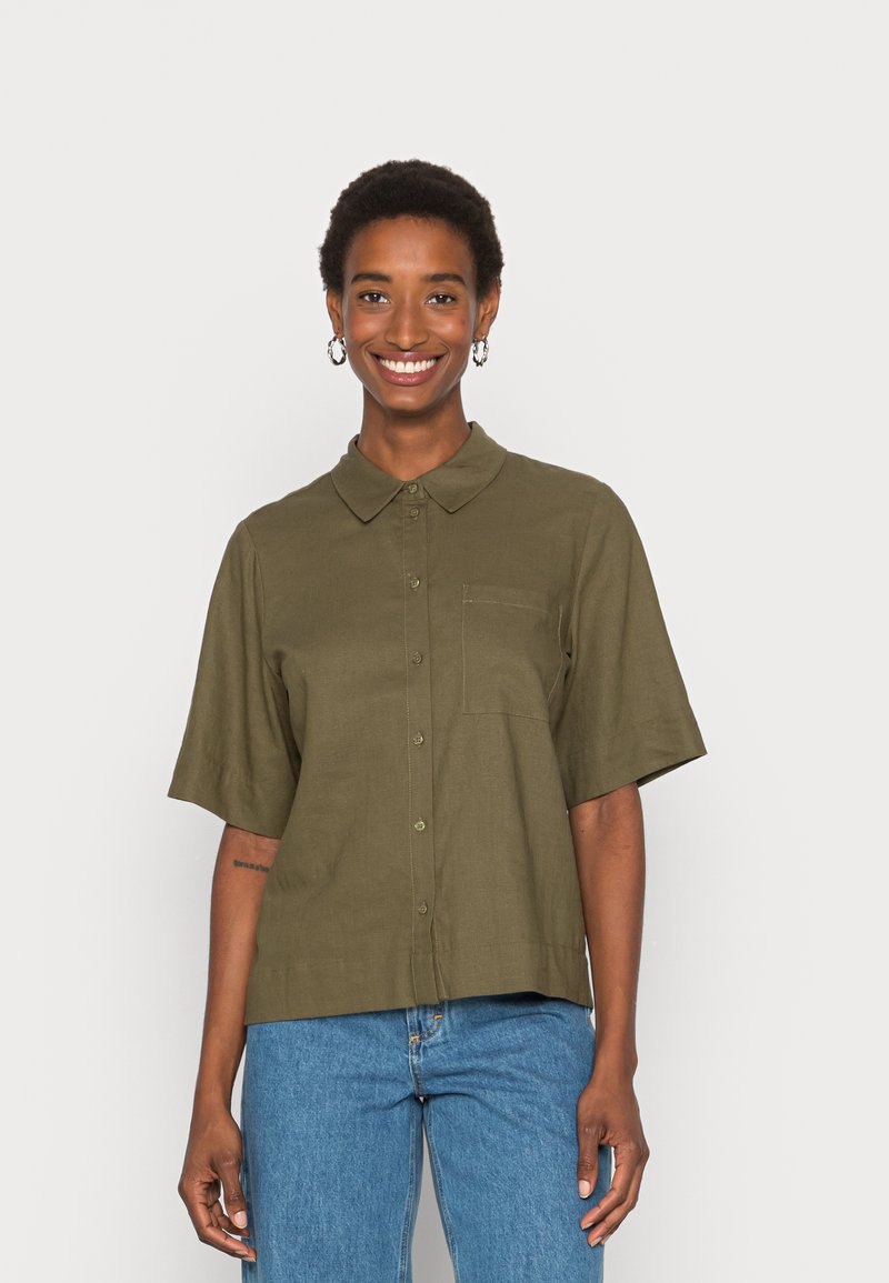 Camisa de manga curta, verde oliva, com fecho de botões à frente, colarinho e um único bolso no peito. Feita de um tecido liso e leve.