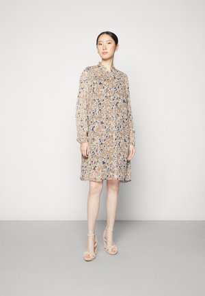 Γυναίκα ντυμένη με μια μπεζ φλοράλ φόρεμα με μακριά μανίκια και κολάρο δεμένο, σε συνδυασμό με μπεζ ψηλοτάκουνες σανδάλες, σε μια γκρι επιφάνεια.