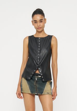 Jonge vrouw draagt een mouwloze zwarte leren top met knopen en een tweekleurige denim minirok met een ritssluiting aan de voorkant.