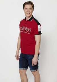 Piros polo, "KRSH" logóval, rövid ujjú, fekete és csíkos részletekkel. Sötét farmer rövidnadrággal viselve, ami egy laza nyári öltözetet mutat.
