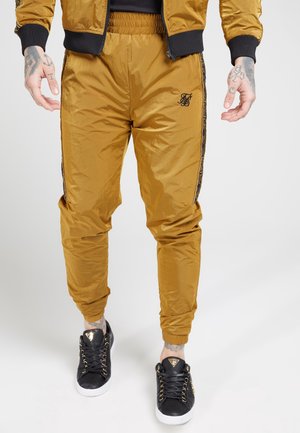 Man draagt okergele trainingsbroek met elastische boorden en zijstrepen, gecombineerd met zwarte sneakers met gouden details.