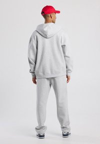 Hombre con una sudadera ligera gris y pantalones de chándal a juego, usando zapatillas navy y blancas, de pie de espaldas sobre un fondo plano.