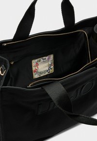 Borsa a tracolla nera in tessuto con una cerniera dorata. L'interno presenta una tasca con motivo floreale e un logo. Manici spessi per un facile trasporto.