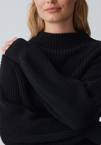 Zwart gebreide trui met een hoge, geribde kraag en losse mouwen, met een gestructureerd patroon en zachte stof.