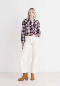Blusa a maniche lunghe a scacchi neri, bianchi e rosa con colletto annodato; abbinata a pantaloni bianchi a vita alta e gamba larga e stivali alla caviglia marroni.