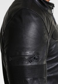 Schwarze Lederjacke mit einem Reißverschlusstaschen an den Ärmeln, geripptem Textur an den Schultern und glatter Oberfläche am Körper.