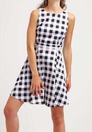 Robe sans manches à carreaux noir et blanc, longueur aux genoux, portée par une personne debout devant un fond uni.