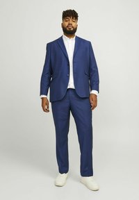 Marineblauer Anzug mit Jacke und passender Hose, aus glattem Stoff. Darunter ein weißes Hemd, getragen mit weißen Sneakern.