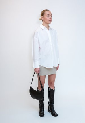 Femeie care stă în picioare, purtând o cămașă albă supradimensionată, fustă mini bej, cizme negre până la genunchi, ținând o geantă neagră pe un fundal simplu.