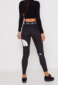 Mustat legginsit korkealla vyötäröllä, joissa on valkoisia logo-aksentteja. Sujuva pinta, tiukka muotoilu ja kaareva graafinen kuvio reidessä.