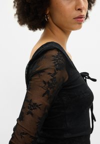 Μαύρη δαντελένια μπλούζα με λουλουδάτα μοτίβα, εφαρμοστή γραμμή και λεπτομέρεια με κόμπο στον λαιμό. Μακρυά, διάφανη μανίκια προσθέτουν υφή και αντίθεση.