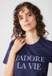 Námořnická modrá bavlněná tričko se textem "J’ADORE LA VIE" v světlejším písmu. Kulatý výstřih a krátké rukávy, hladká textura.