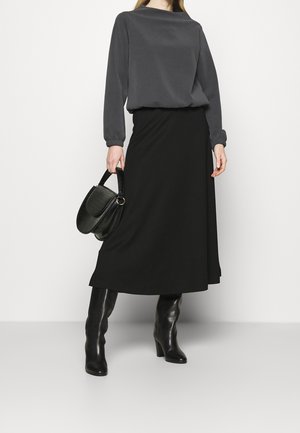 Kobieta w długiej czarnej spódnicy, ciemnoszarej bluzce z długim rękawem, czarnych kozakach do kolan, trzymająca czarną teksturowaną torebkę z krótką rączką.