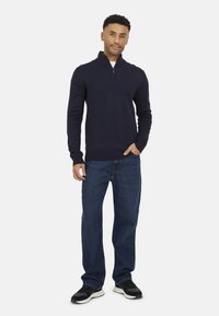 Maglione navy a manica lunga con zip a quarti, dal design aderente, abbinato a jeans in denim blu larghi e scarpe da ginnastica nere con suola bianca.
