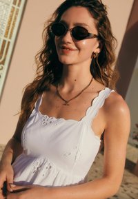 Vestido de algodón blanco con patrones florales bordados, tirantes ondulados y una silueta acampanada. Combinado con joyería dorada y gafas de sol.