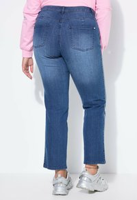 Persona che indossa jeans di jeans blu, maglietta a maniche lunghe rosa e scarpe sportive argento, in piedi su sfondo bianco.