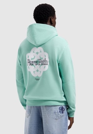 Man draagt een lichtgroene hoodie met een wit decoratief logo en de tekst "Pure Path" op de achterkant, gecombineerd met lichtblauwe spijkerbroek.