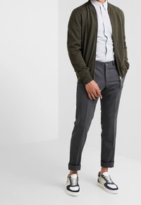 Olivgrüner Strickbomberjacke über einem hellblauen Hemd mit Knöpfen, kombiniert mit maßgeschneiderten grauen Hosen und mehrfarbigen Sneakers.