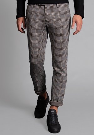 Mann trägt hochgekrempelte graue Karottenhose und schwarze Turnschuhe, geht vor einem schlichten grauen Hintergrund.