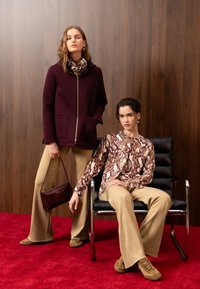 Deux femmes en pantalon beige et chaussures marron, l'une debout avec une veste bordeaux et un sac à main, l'autre assise sur une chaise noire portant un haut à imprimé serpent.