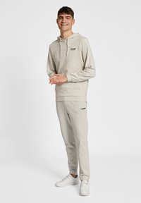 Helles graues Hoodie und Jogginghose aus weichem Stoff mit Rippbündchen. Mit kleinem schwarzen Logo auf der Brust und Hüfte. Getragen mit weißen Sneakern.