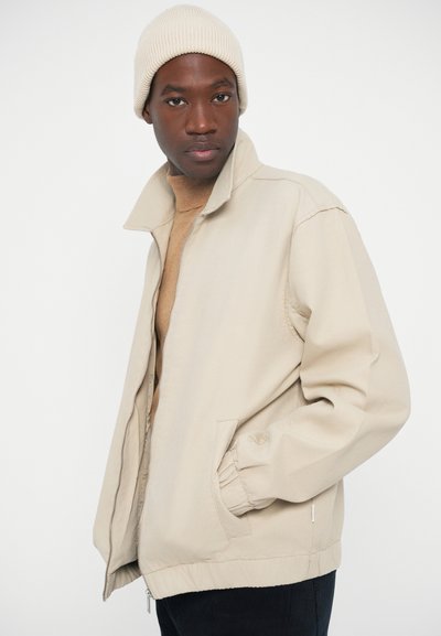 Beige Kapuzenjacke mit Reißverschluss, Kragen, elastischen Bündchen und Seitentaschen. Getragen über einem braunen Strick-Oberteil, kombiniert mit einer beigen Mütze.