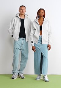Sweats à capuche gris avec zip intégral, associés à un jean en denim bleu clair. Le modèle féminin porte une jupe fendue ; le modèle masculin porte un jean décontracté.