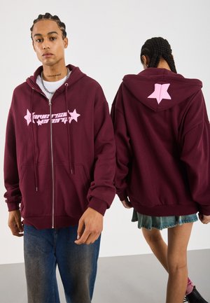Two people wearing maroon zip-up hoodies with pink star designs; one faces front, the other shows the hood's back with a pink star.