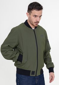 Zielona oliwkowa kurtka bomberka z czarnym prążkowanym kołnierzem i mankietami, zamek z przodu, kieszenie boczne i kieszeń na rękawie z zamkiem błyskawicznym na drobiazgi.