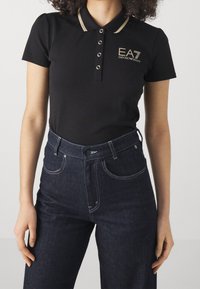 Svart polotröja med en guld-accentuerad krage, tre knappar och broderad EA7-logotyp; ihop med mörka högmidjade jeans.