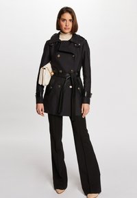Svart trench coat med dobbeltkneppdesign, sølvknapper og et strukturert svart belte. Kombinert med vide bukser og en hvit håndveske.