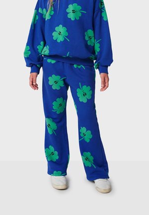 Blue sweatshirt and matching pants, both featuring large green motifs of clovers with black accents, made of soft material. White shoes visible.