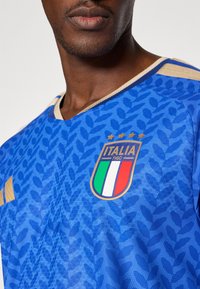 Plava nogometna majica s teksturiranim uzorkom, dizajnom u obliku slova V, zlatnim detaljima i oznakom nacionalne momčadi Italije s obojenim prugama.