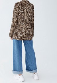 Blazer à imprimé léopard en tissu doux, associé à un jean denim à jambes larges, ourlets retroussés, et baskets blanches. Vue de dos sur un fond uni.