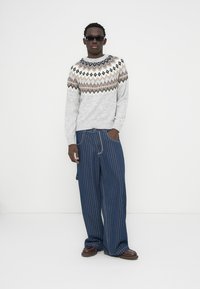 Pull en laine gris avec un motif géométrique brun et noir au niveau de l'encolure, associé à un pantalon large bleu à fines rayures. Le mannequin porte des lunettes de soleil.