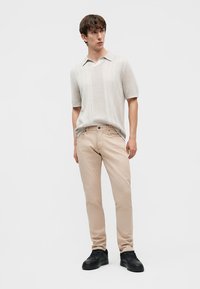 Hombre joven de pie, con una camisa beige de manga corta y textura, pantalones de color beige claro y zapatillas negras, sobre un fondo liso y claro.