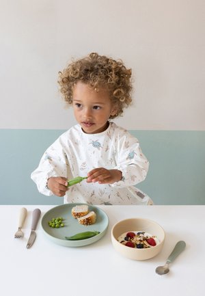 Unge barn med krøllet hår, der skaller ærter ved bordet med en tallerken brød, ærter og en skål yoghurt med bær og granola.