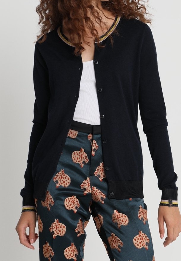 Marinefarbener Cardigan mit Rundhalsausschnitt, kontrastierendem gelbem und schwarzem Rippstricksaum und Knopfverschluss, kombiniert mit gemusterten Hosen, die Tiermotive zeigen.