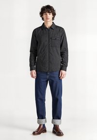 Sort quiltet skjortejakke med frontlynlås og brystlomme, parret med mørkeblå opsmøgede jeans og brune læderstøvler. Glat tekstur.