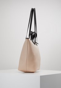 Torebka tote w kolorze brzoskwiniowym, o zarysowanej formie, z czarnymi skórzanymi ramiączkami i gładką fakturą. Minimalistyczny design bez widocznych elementów metalowych.