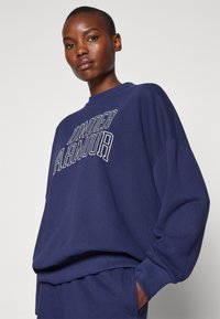 Kvinde med kort hår iført en navy blue Under Armour sweatshirt og matchende bukser, står foran en ensfarvet lys baggrund.