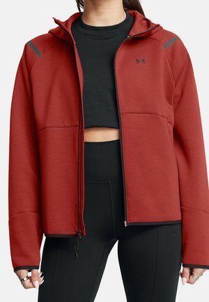 Kvinde iført en rød zip-up hættetrøje over en sort kort top og sorte højtaljede leggings, med ringe på fingrene.