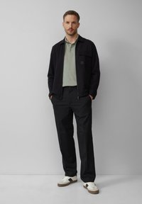 Schwarze Zip-Jacke über einem hellgrünen Poloshirt, kombiniert mit schwarzen Hosen und weißen Sneakers mit braunen Akzenten; flache Textur und modern geschnitten.