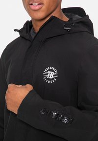 Schwarze Fitnessjacke mit Reißverschluss an der Kapuze, strukturiertem Stoff und einem runden Logo auf der Brust. Reflektierende Akzente am Ärmel.