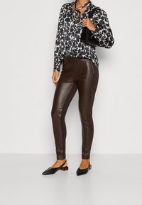 Mujer vestida con blusa de estampado en blanco y negro, pantalones de cuero marrón, zapatos de tacón negro tipo slingback y llevando un bolso negro al hombro.