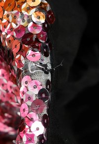 Gros plan d'un tissu recouvert de sequins ronds brillants roses et orange cousus sur une matière en maille noire et grise.