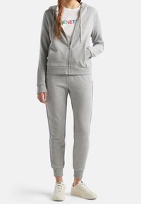 Sudadera gris con cremallera y pantalones de chándal a juego, ambos de tela suave. La sudadera tiene cordones y puños acanalados; logo mostrado en la camiseta de abajo.