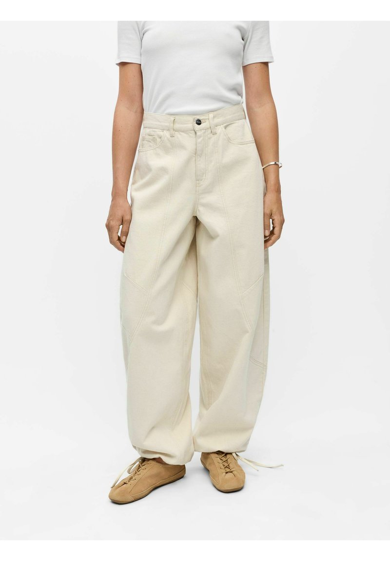 Personne portant un pantalon beige taille haute à jambes larges avec des détails de couture et des chaussures à lacets marron, debout devant un fond blanc uni.