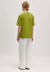 Olivgrünes T-Shirt mit lässigem Schnitt, kurzen Ärmeln und Rundhalsausschnitt, kombiniert mit weiten weißen Hosen und schwarzen Schuhen.